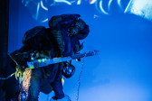 A performer wears a suit adorned with large appendages and plays a stringed instrument. The stage and wall behind it are bathed in blue light. 