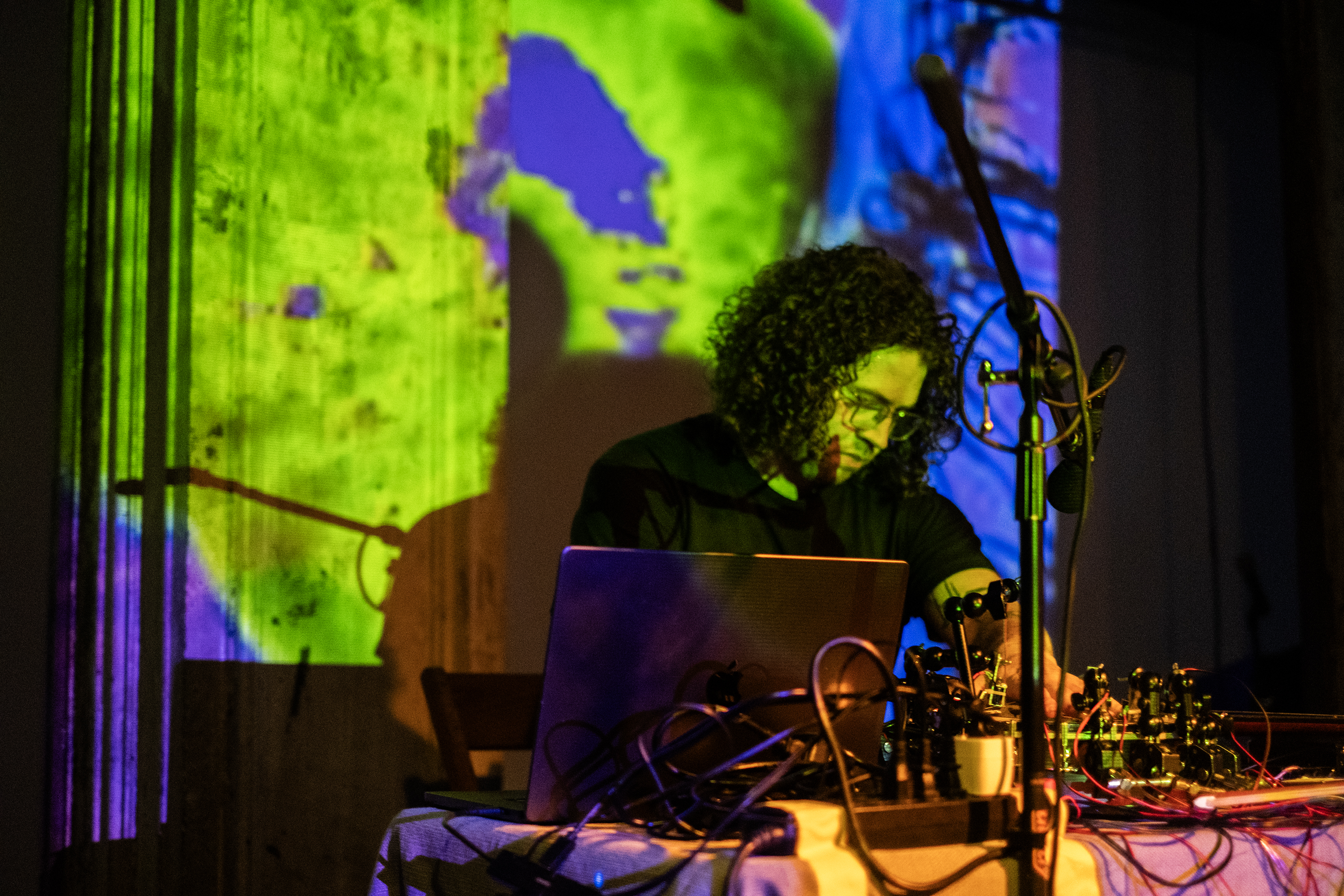 A performer is surrounded by equipment, playing on stage. The stage is bathed in green and blue light patterns created by a projection.