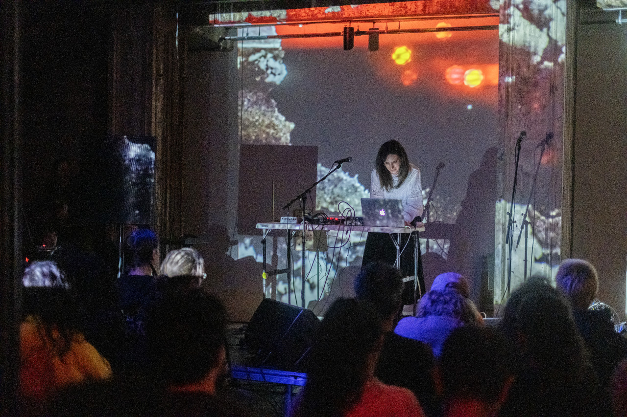 A performer stands, engaged with a sound system on stage in front of an audience. The stage and wall behind the stage are bathed in iridescent light produced by a projection.