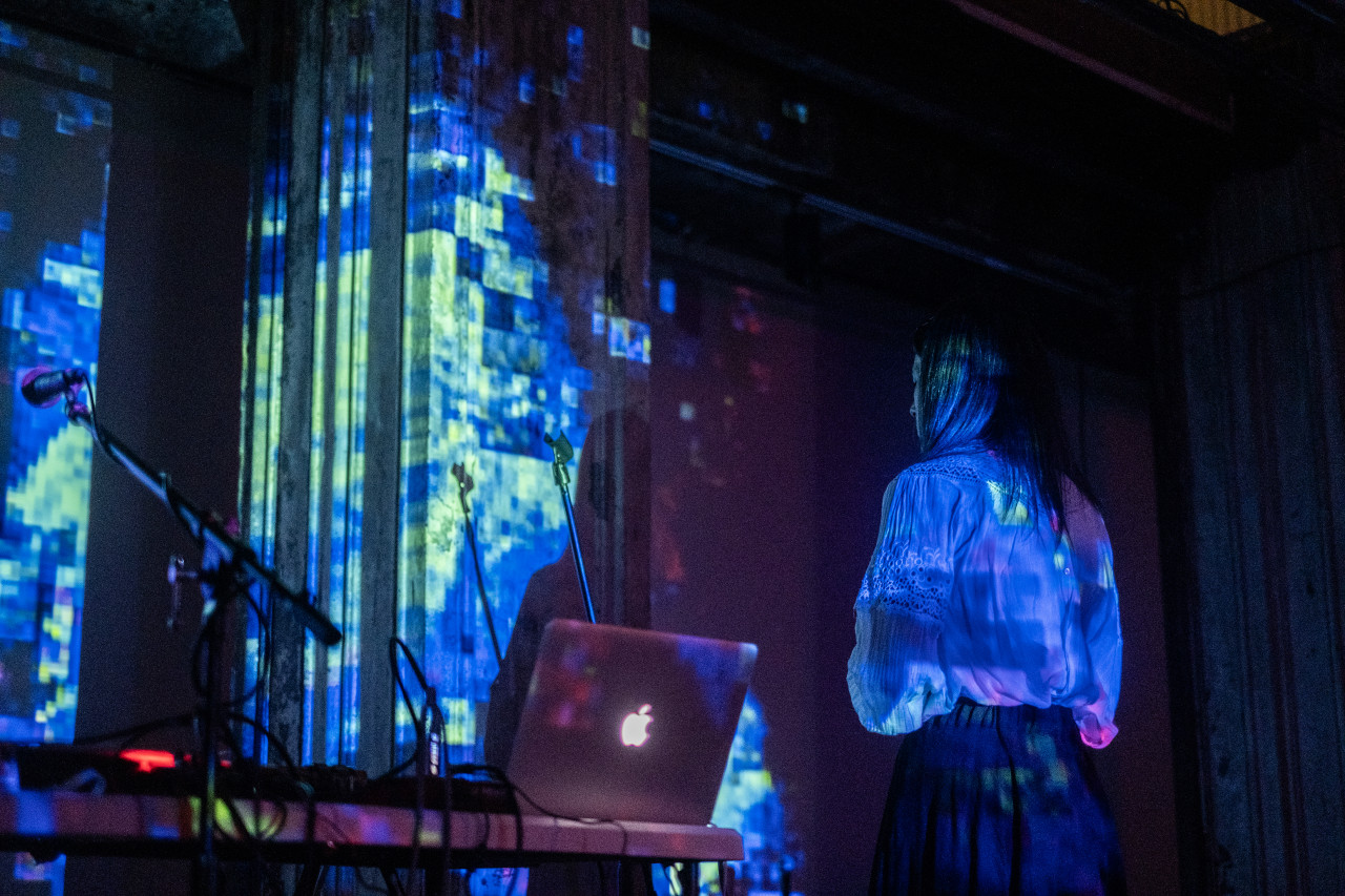 A performer on stage faces the wall. Both the performer and wall are bathed in speckled blue and green lights.