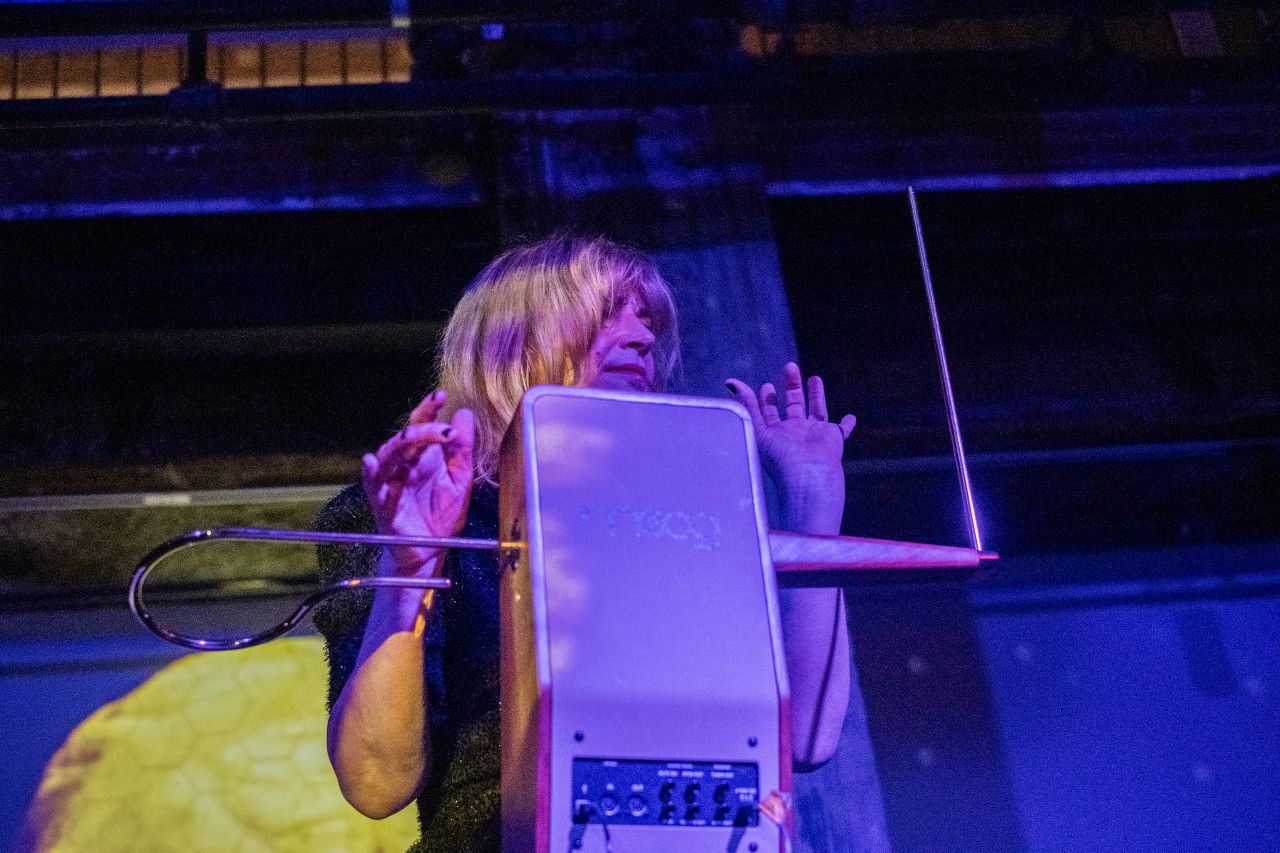 A performer plays the theremin on stage, bathed in pink and blue light.