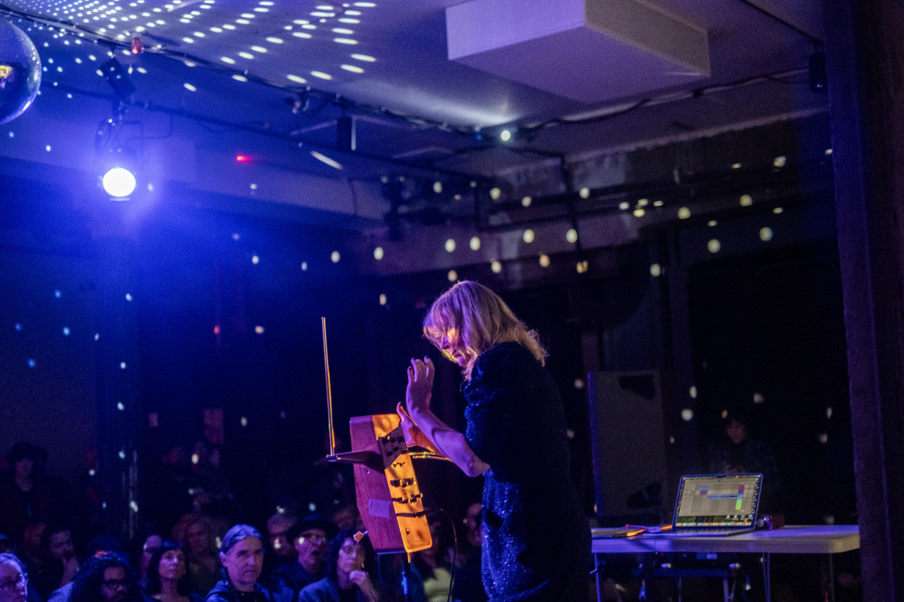 A performer facing an audience plays the theremin on stage. The performer is lit by a spotlight, and the room is bathed in a blue and twinkling white light.