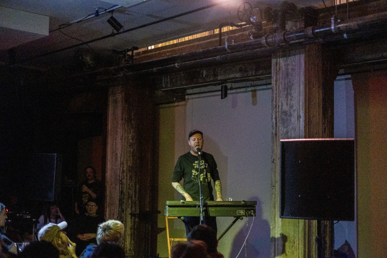 A performer sings and plays the keyboard on stage, facing an audience. The stage is lit with a yellow light.