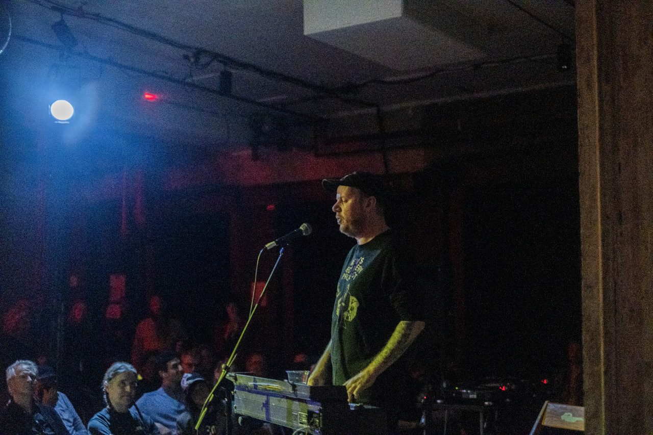 A performer sings and plays the keyboard on stage, facing an audience. The performer is lit by a spotlight, and the audience is bathed in a red and blue light.