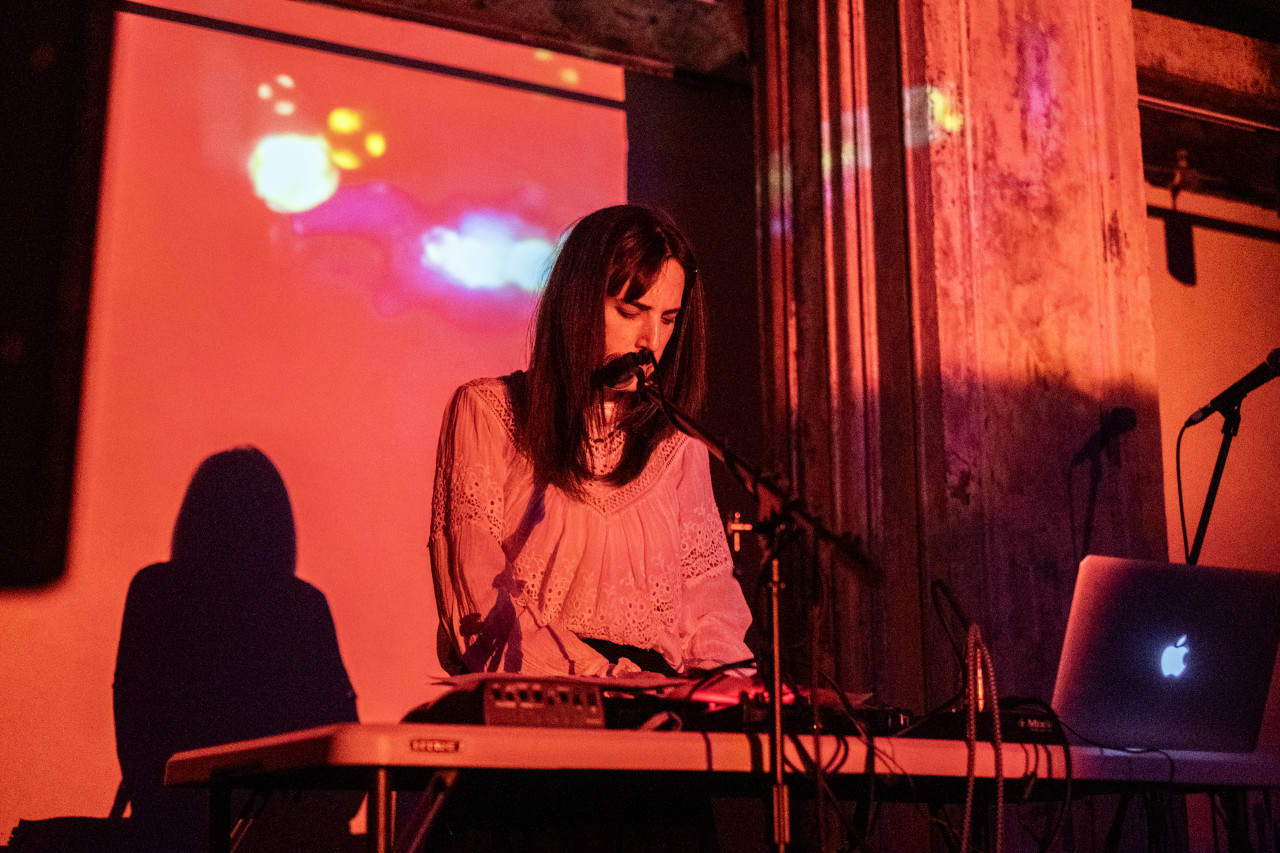 A performer stands on stage, operating a sound system. The performer and the wall behind the stage are illuminated in red light.
