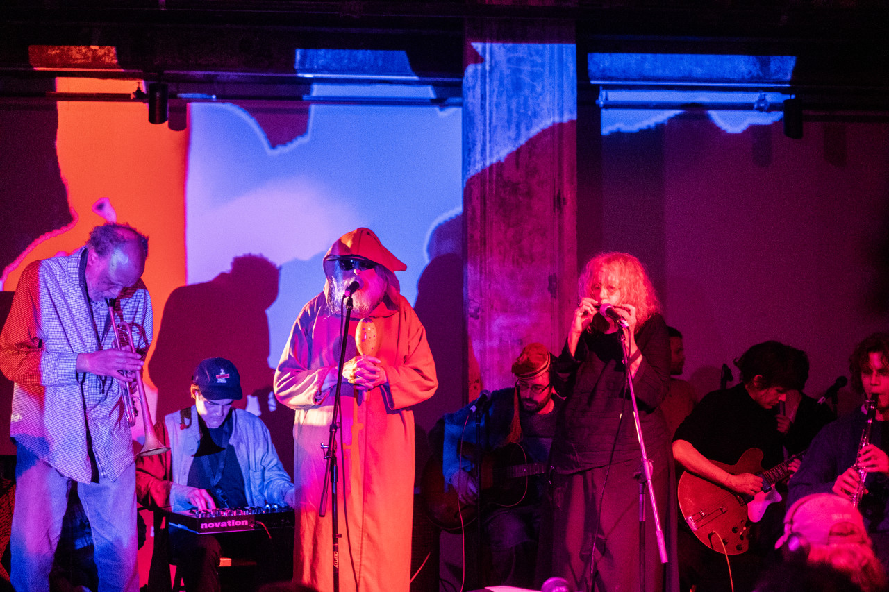 A group of performers plays various instruments on stage while bathed in pink, red, and blue light.