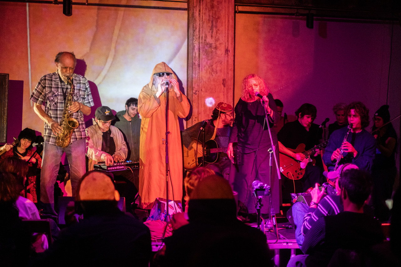 Twelve performers play various instruments on stage for an audience. The stage and performers are bathed in red and orange light. A member sitting in the front row videotapes the performance.