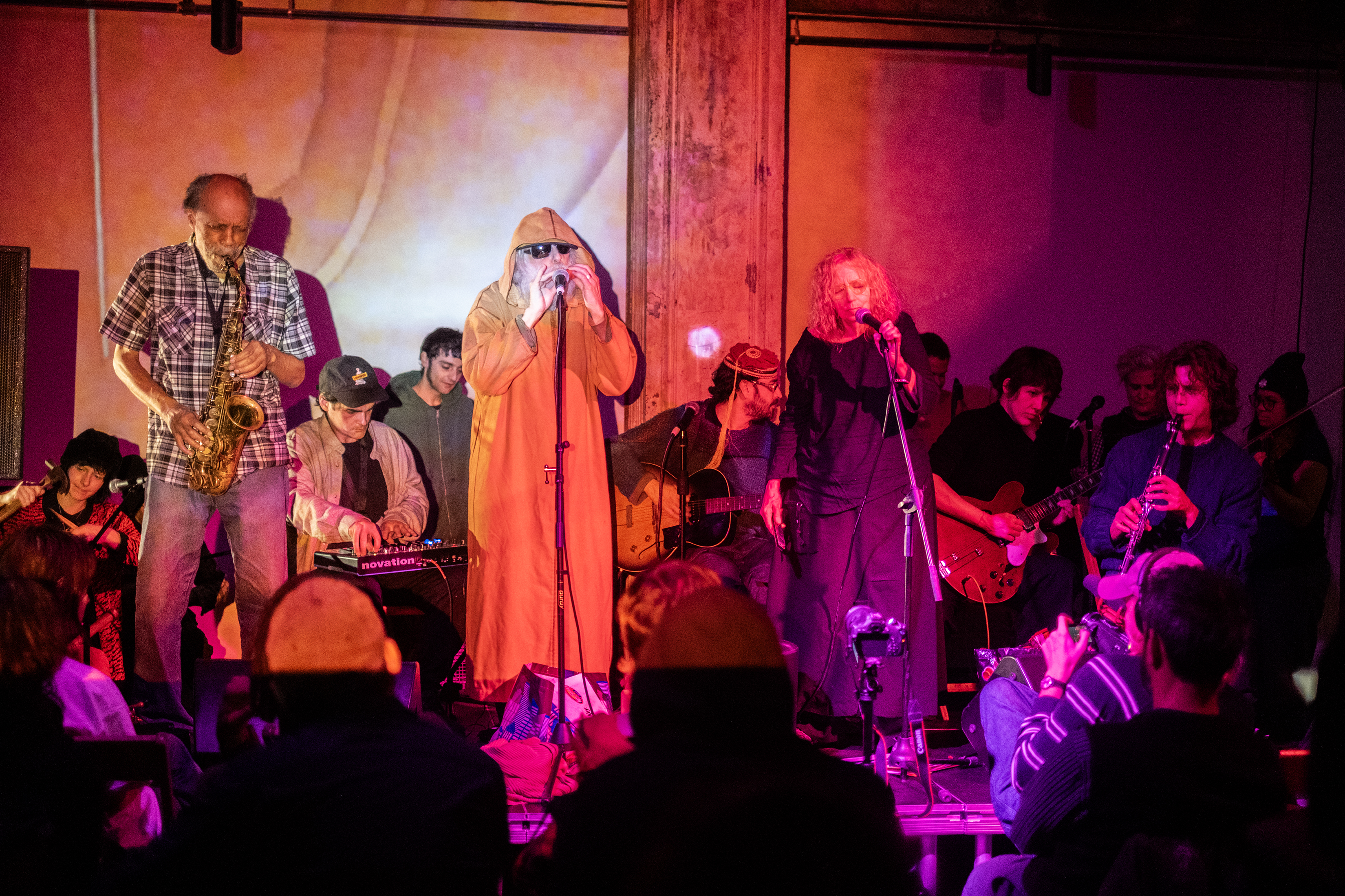 Twelve performers play various instruments on stage for an audience. The stage and performers are bathed in red and orange light. A member sitting in the front row videotapes the performance.