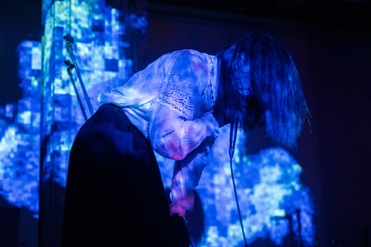 A performer sings onstage, obscured by hair and bathed in shades of blue light.