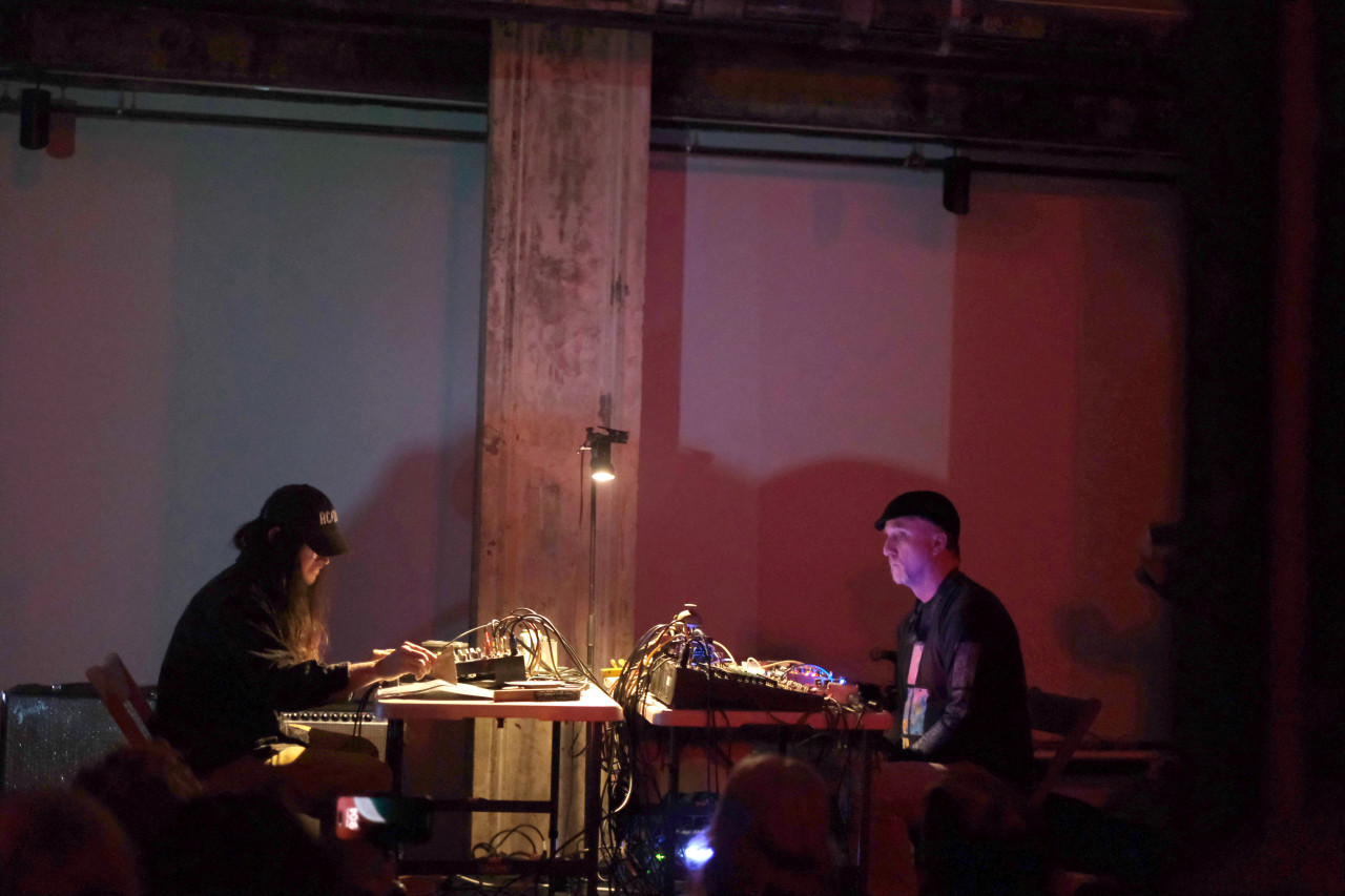 Two performers sit at tables facing eachother on stage. Each performer operates a mixing board abundant with cables. The tables are lit from a single bulb above.