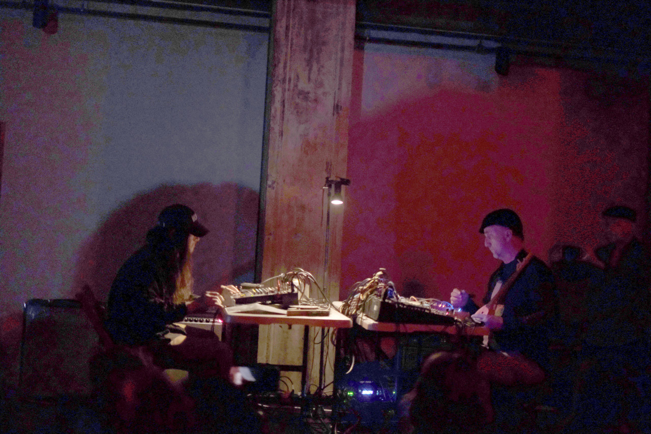 The stage is dark except for a red glow, and a single bulb illuminating two performers sitting at tables facing each other on stage. Each performer operates a mixing board abundant with cables.
