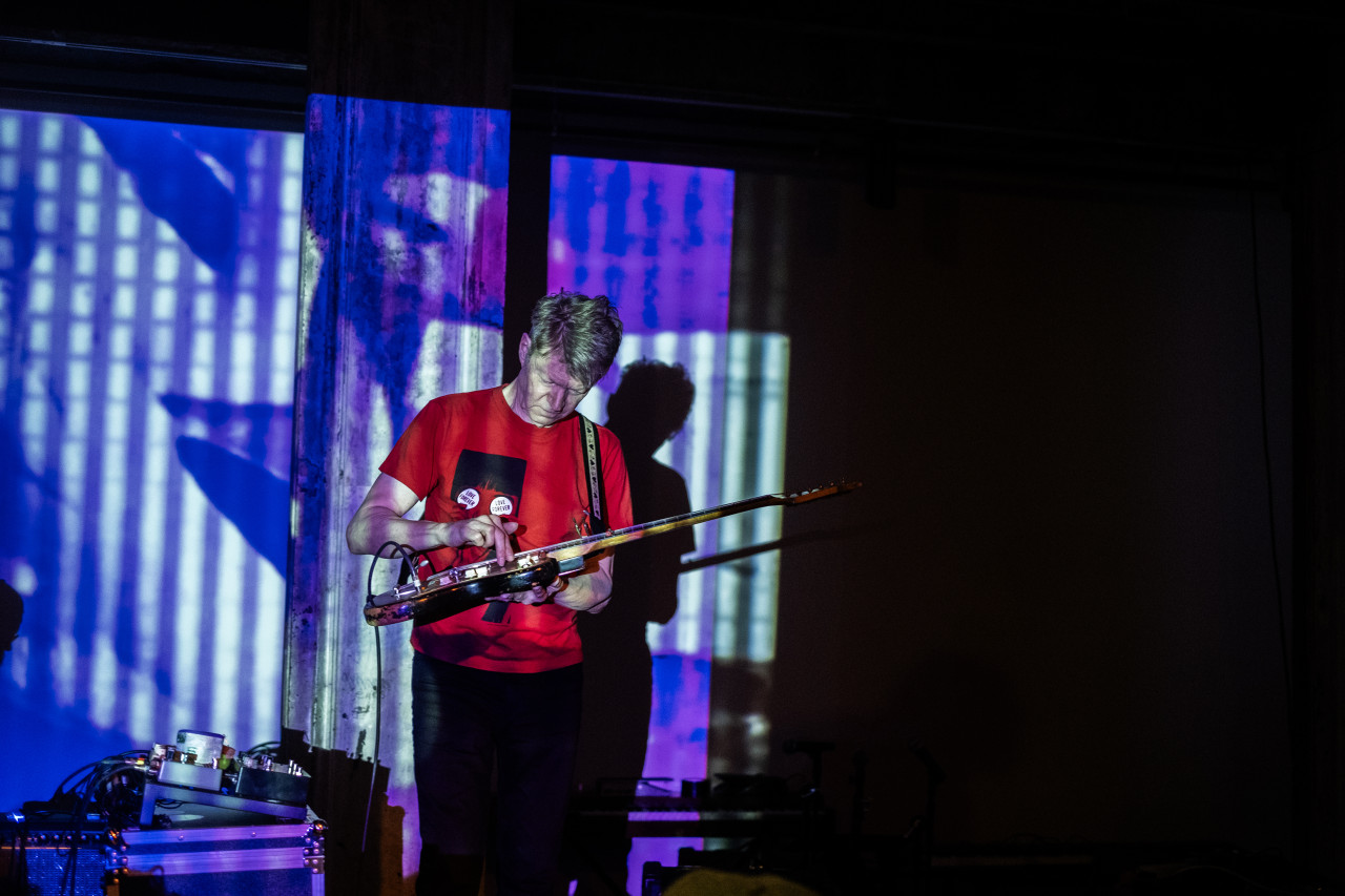 A performer plays an electric guitar on stage. The performer and stage are bathed in blue light and geometric patterns.