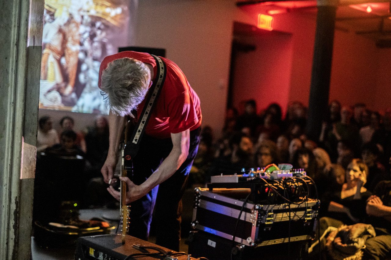 An onlooking audience watches a performer play an electric guitar on stage. The audience is bathed in red light, and projections are present on the left wall.