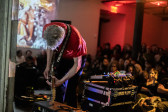 An onlooking audience watches a performer play an electric guitar on stage. The audience is bathed in red light, and projections are present on the left wall. 