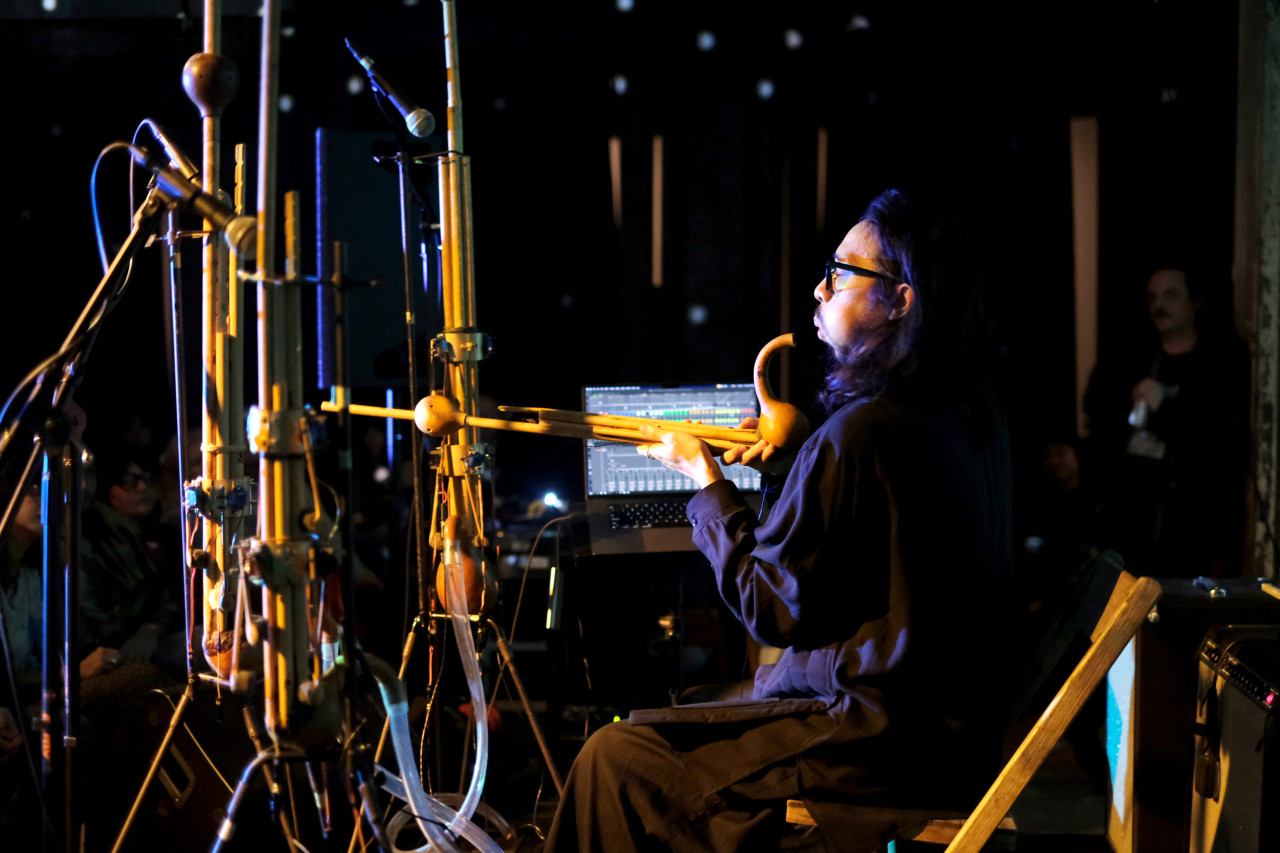 A performer plays a wooden wind instrument on stage. Connected by plastic tubing and wooden rods, the instrument stands higher than the seated performer.