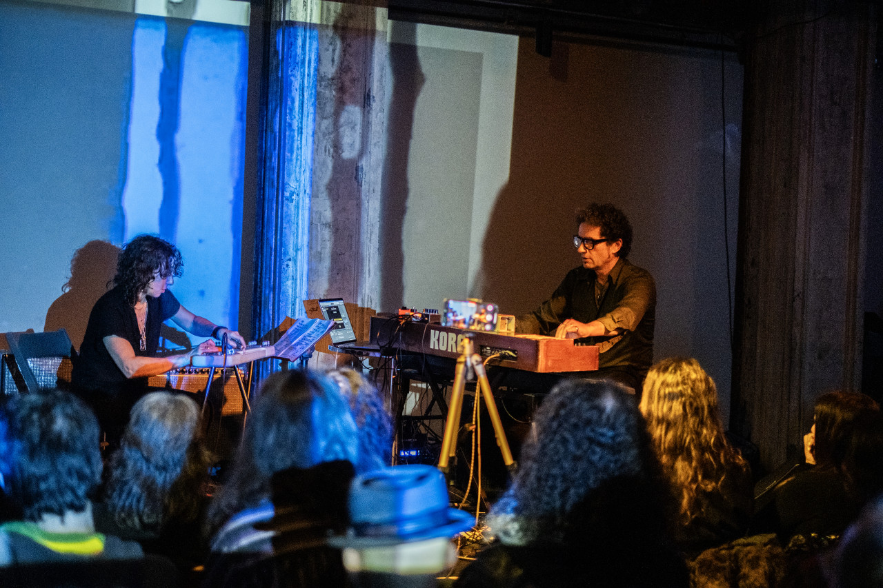 An audience observes two performers playing a keyboard and a stringed instrument on stage. The performers are seated at an angle towards each other, and the stage is bathed in blue and yellow light.