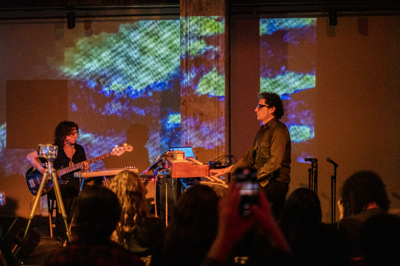 An audience observes two performers playing a keyboard and a bass on stage. The performers are seated at an angle towards each other, and the stage is bathed in orange light and blue cloud-like patterns.