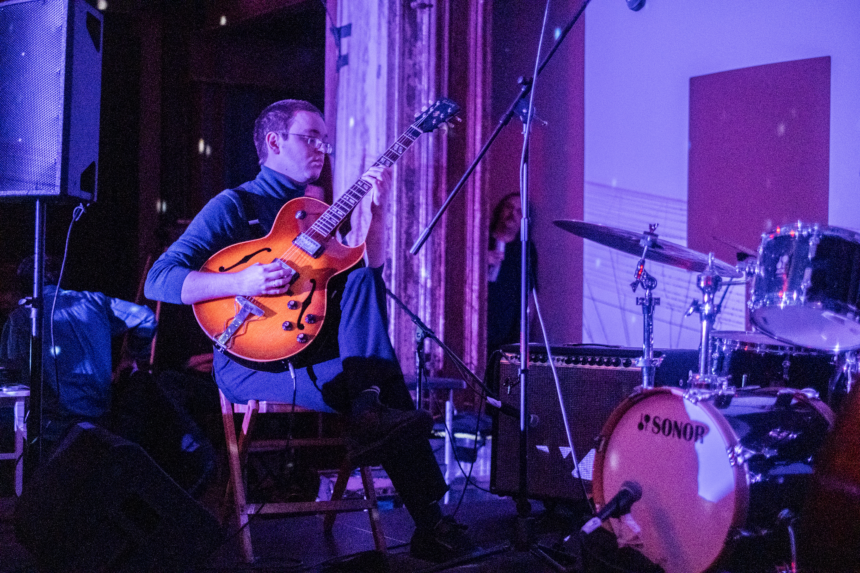 A performer, bathed in purple light, plays a guitar on stage.