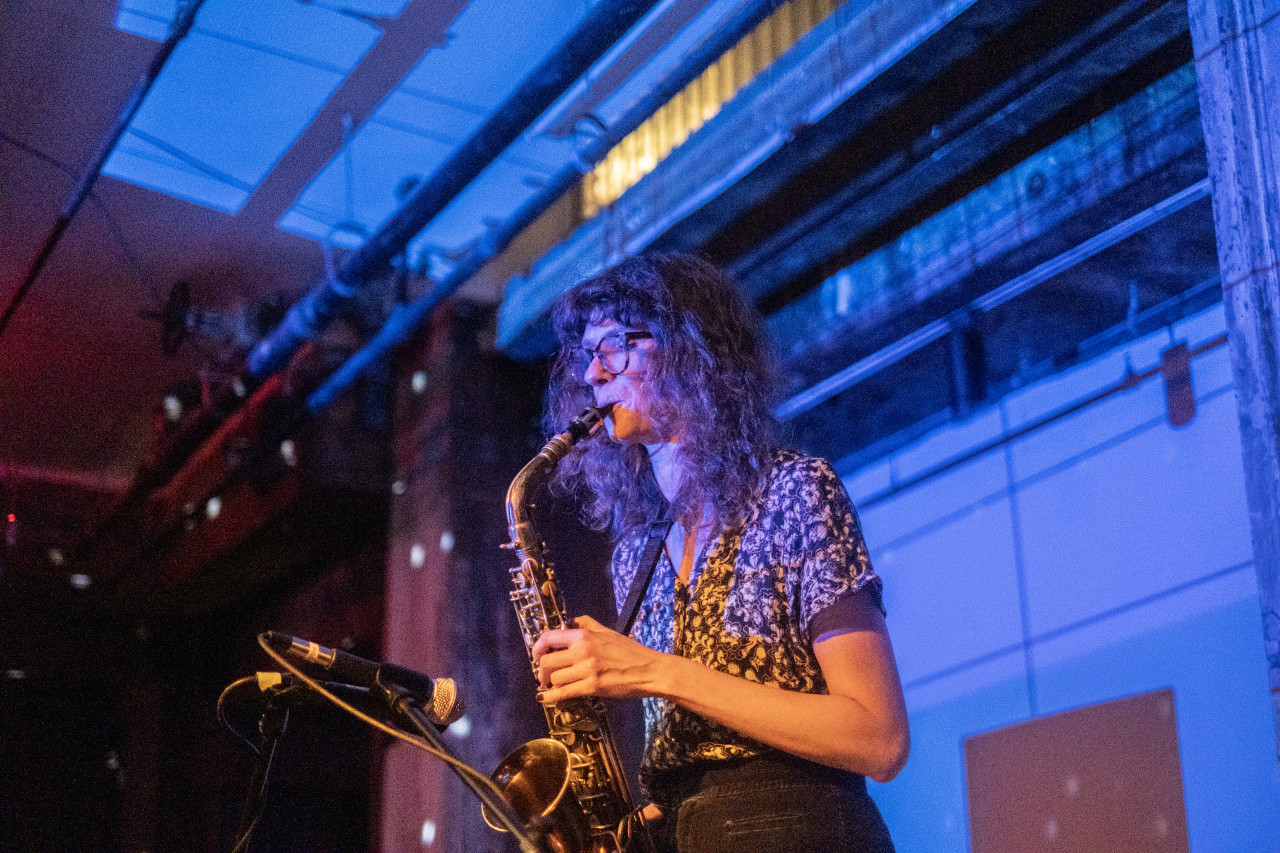 A performer plays the saxophone into a microphone on stage. The wall behind the stage is bathed in blue light.