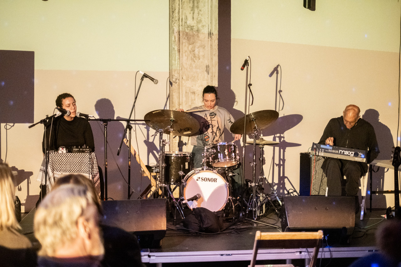 Three performers, from left to right, sit singing, playing the drums, and operating a sound board on stage.