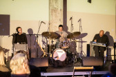 Three performers, from left to right, sit singing, playing the drums, and operating a sound board on stage. 