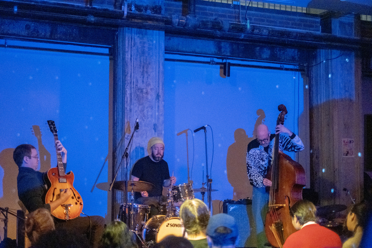 From left to right, three performers play a guitar, the drums, and a standing bass on stage in front of an audience. The stage is illuminated with blue light.