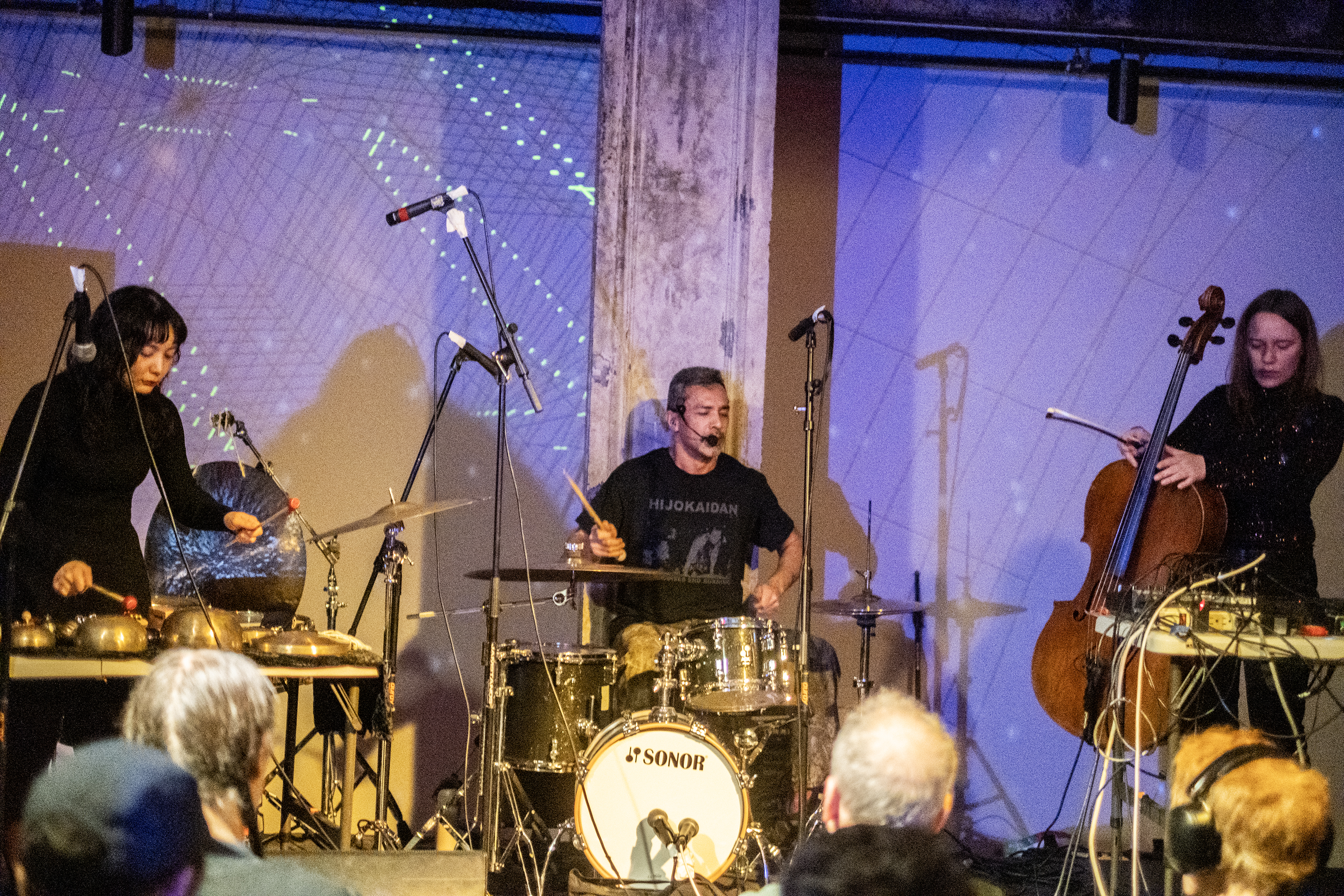 Three performers from left to right play cymbals, drums, and a cello on stage. The wall behind the stage is lit with blue and white lights. 
