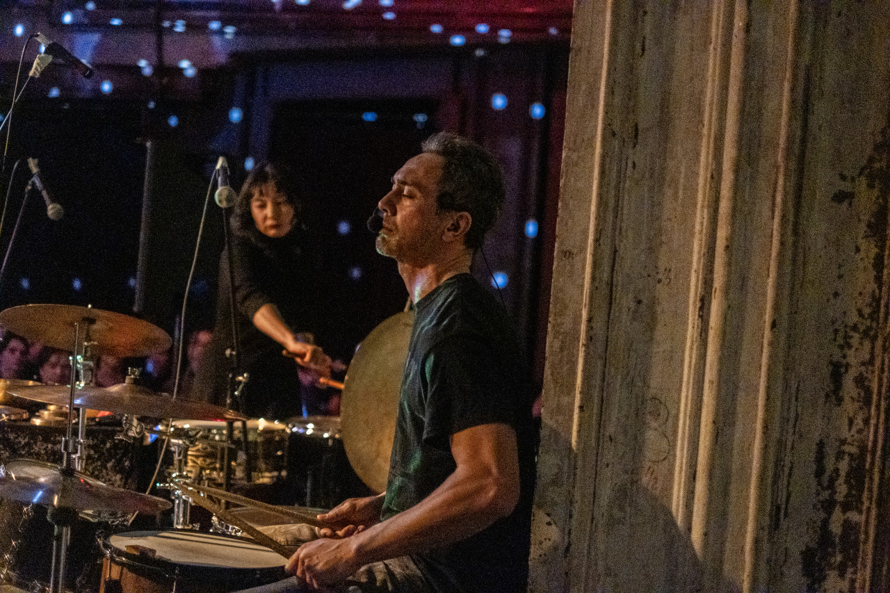 Two performers play the drums on stage. The room is speckled with white dotted lights.