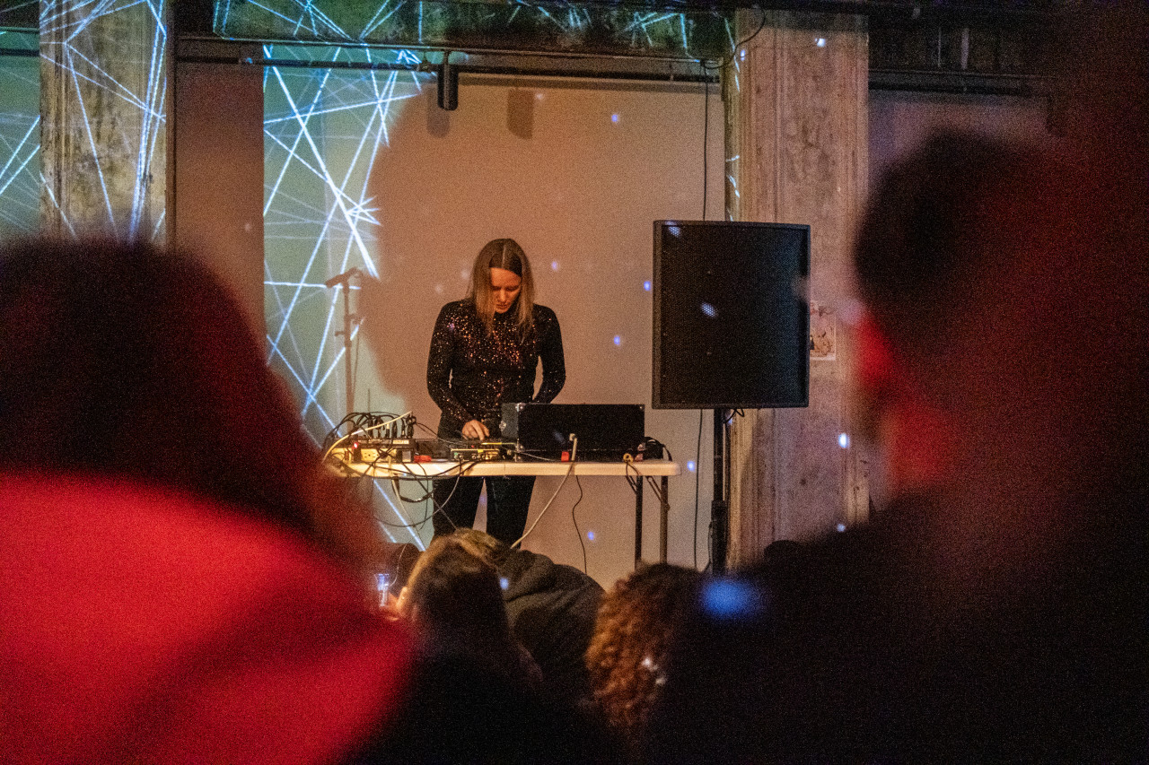 A performer stands behind a table operating a sound system in front of an audience. The wall behind the stage is lit with white geometric patterns.
