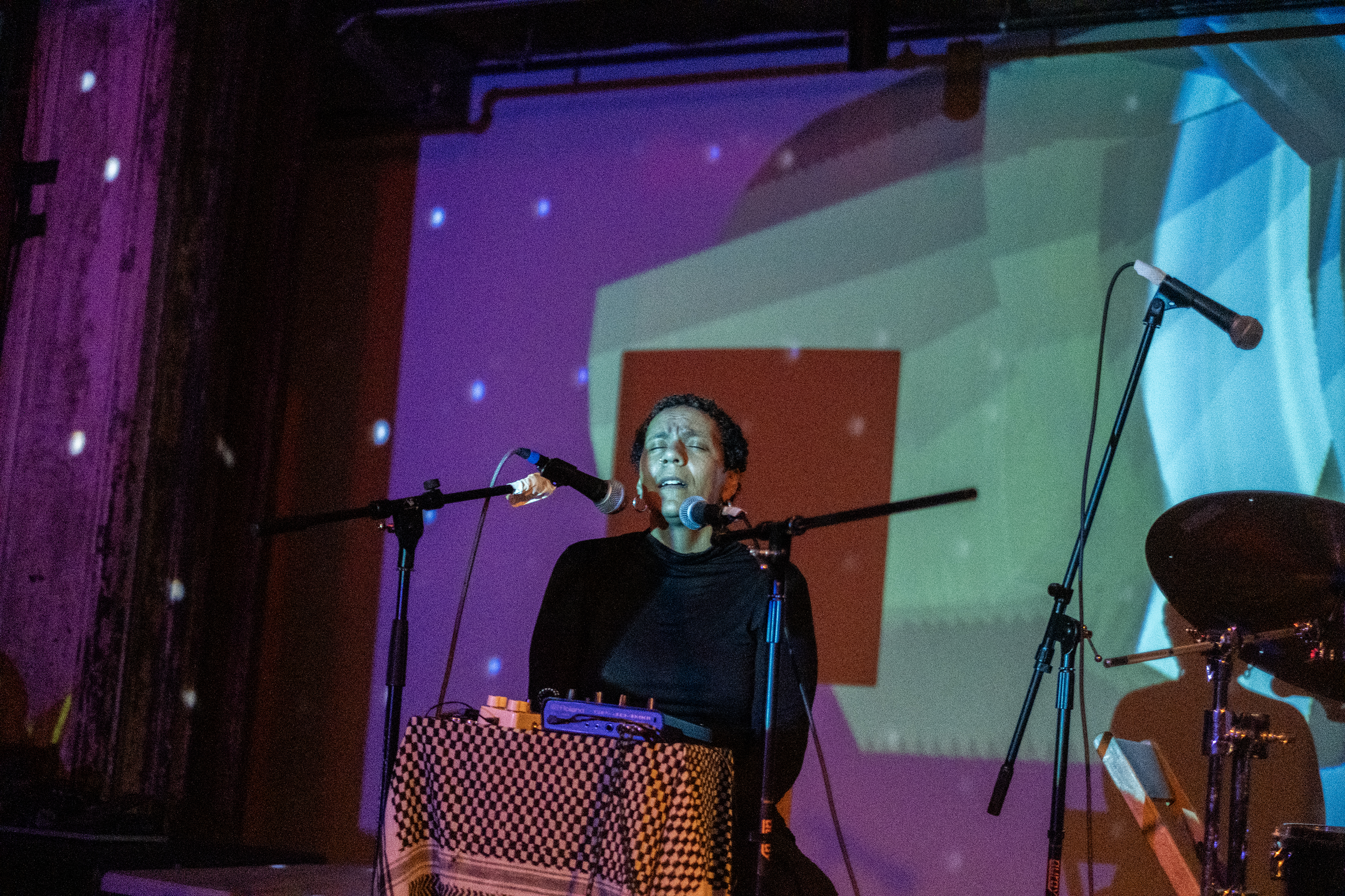 A performer sings into two microphones while green, purple, and white lights illuminate the stage and wall behind it. 