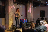 A performer plays the saxophone into a microphone on stage for an audience. Speckled white lights appear around the room. 