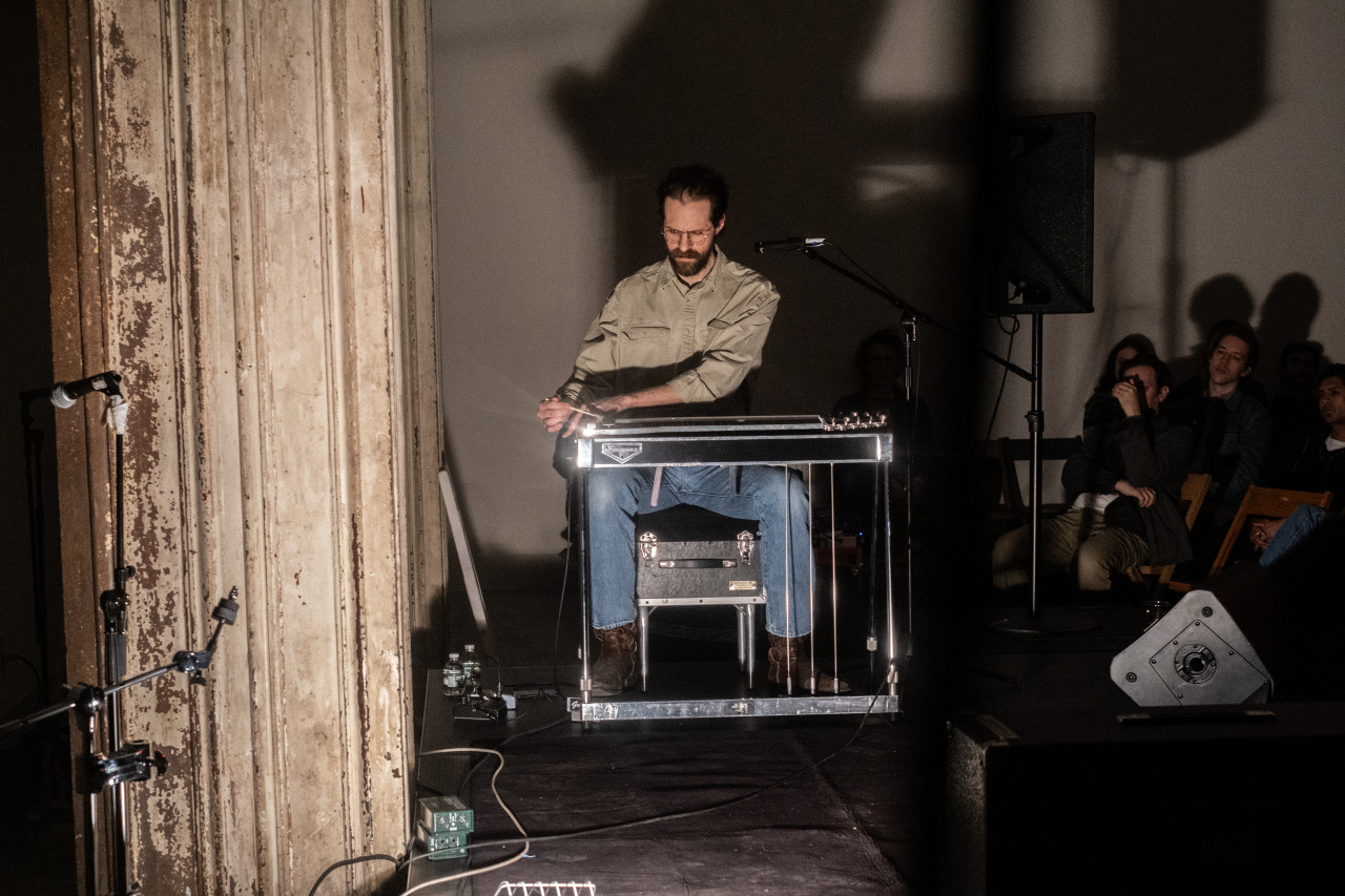 A performer plays a stringed instrument on stage. The performer and the onlooking audience are illuminated in bright lights.