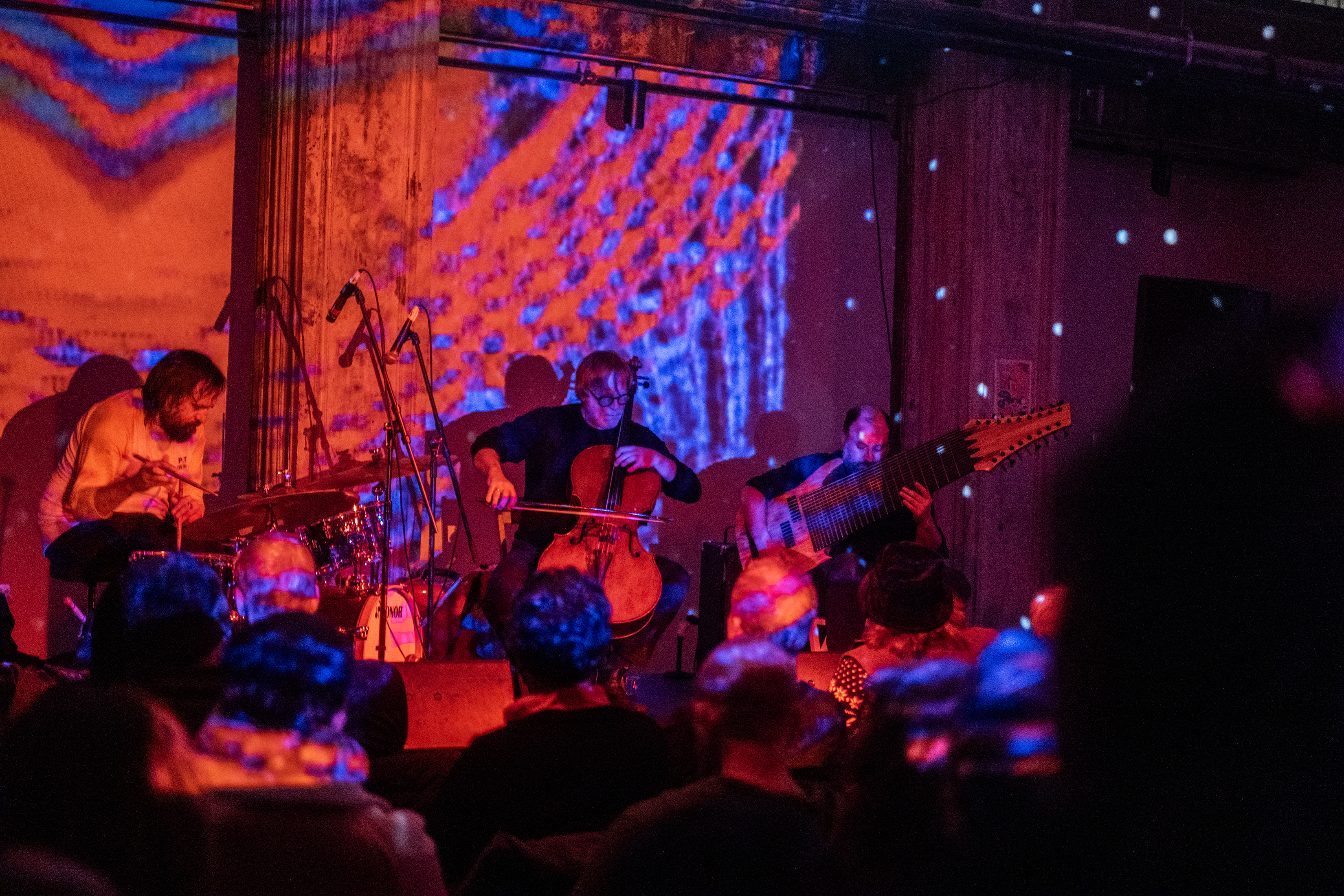 Three performers play various instruments on stage in front of an audience. The stage and wall behind it are bathed in a blue and red striped pattern. 