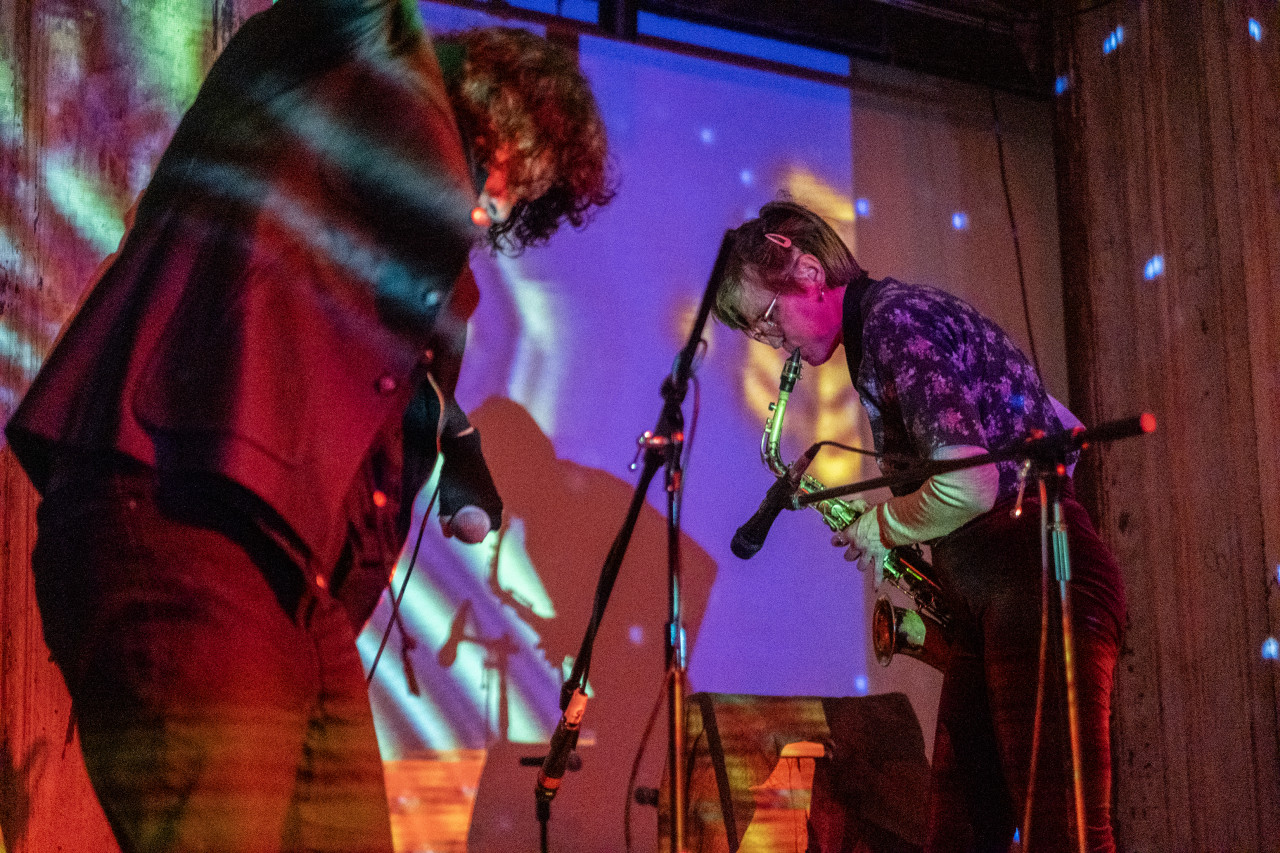 Two performers stand and move on stage. On the right, a performer is playing the saxophone. The stage is illuminated in blue and white lights, as well as a colorful, patterned projection.