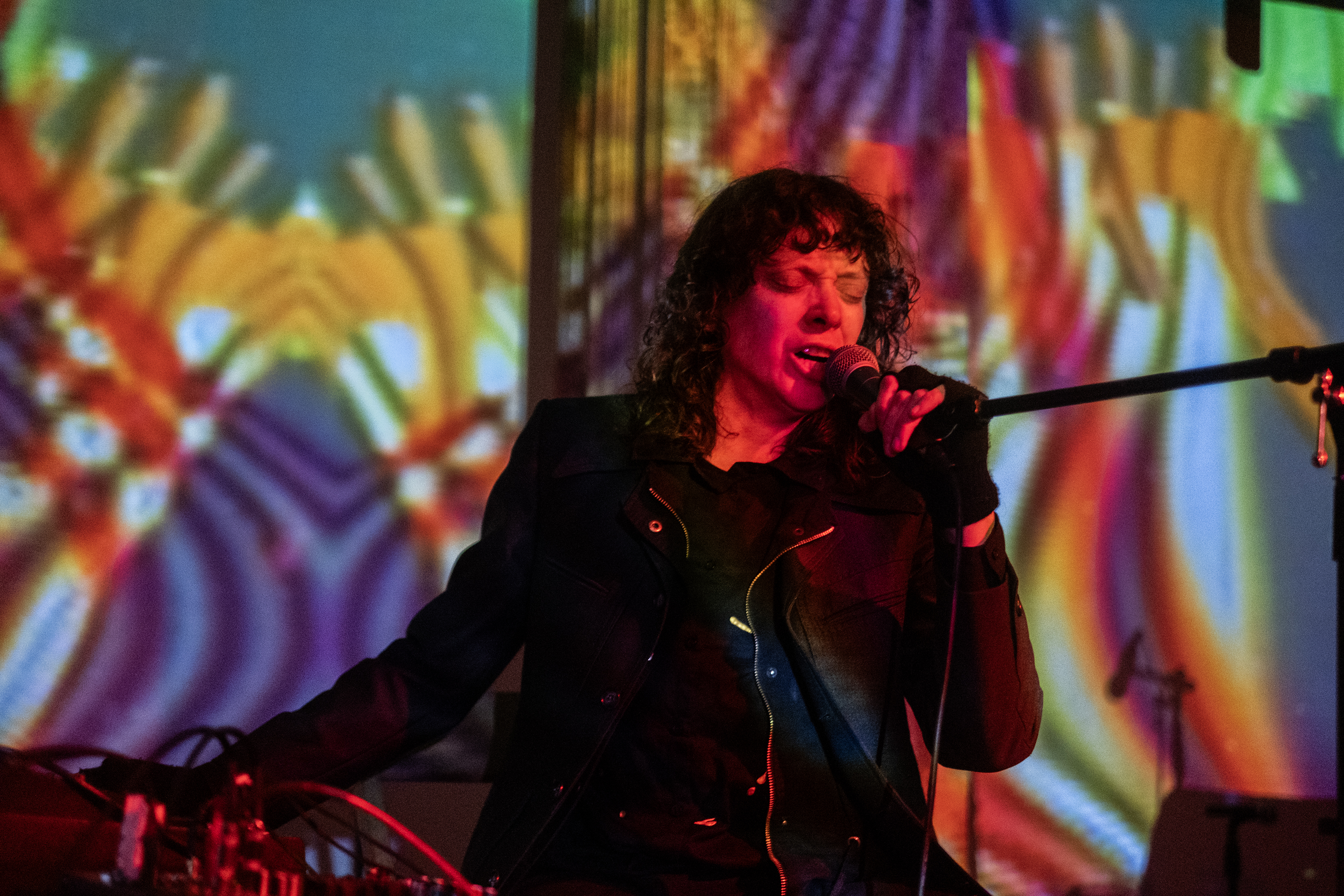 A performer sings into a microphone. The performer and stage are bathed in psychedelic, colorful patterns. 