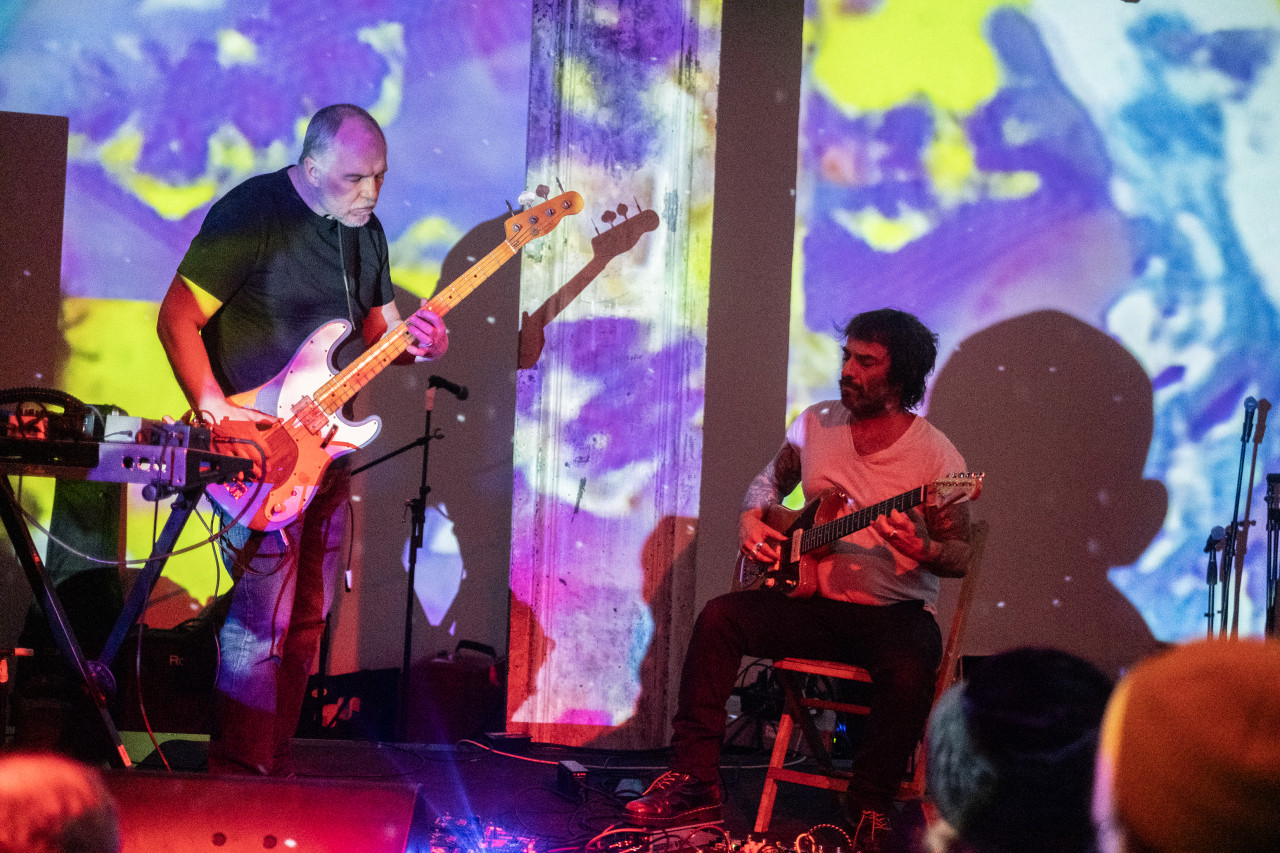 Two performers play an electric bass and an electric guitar. They are bathed in pink light while colorful projections light the wall behind them.