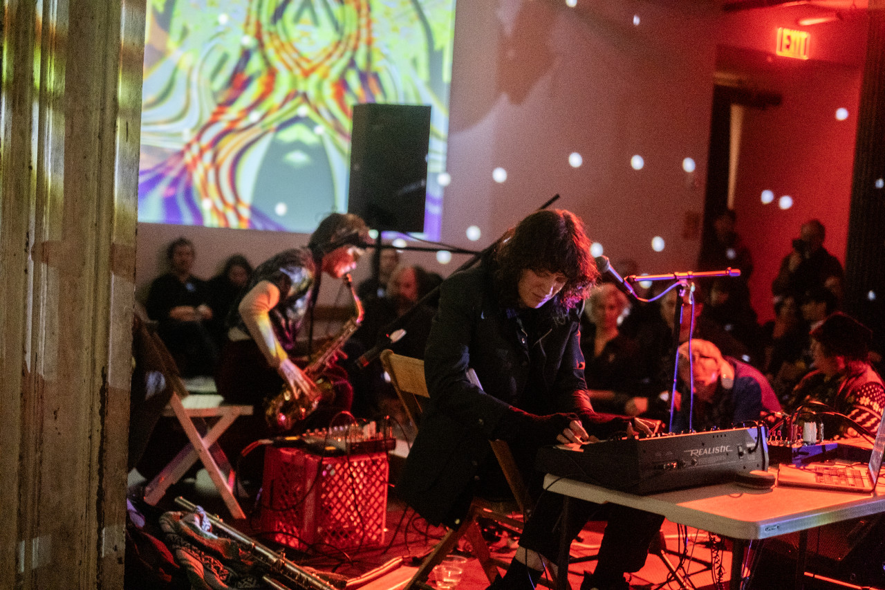 One performer operates a sound system, while the other plays the saxophone on stage in front of an audience.