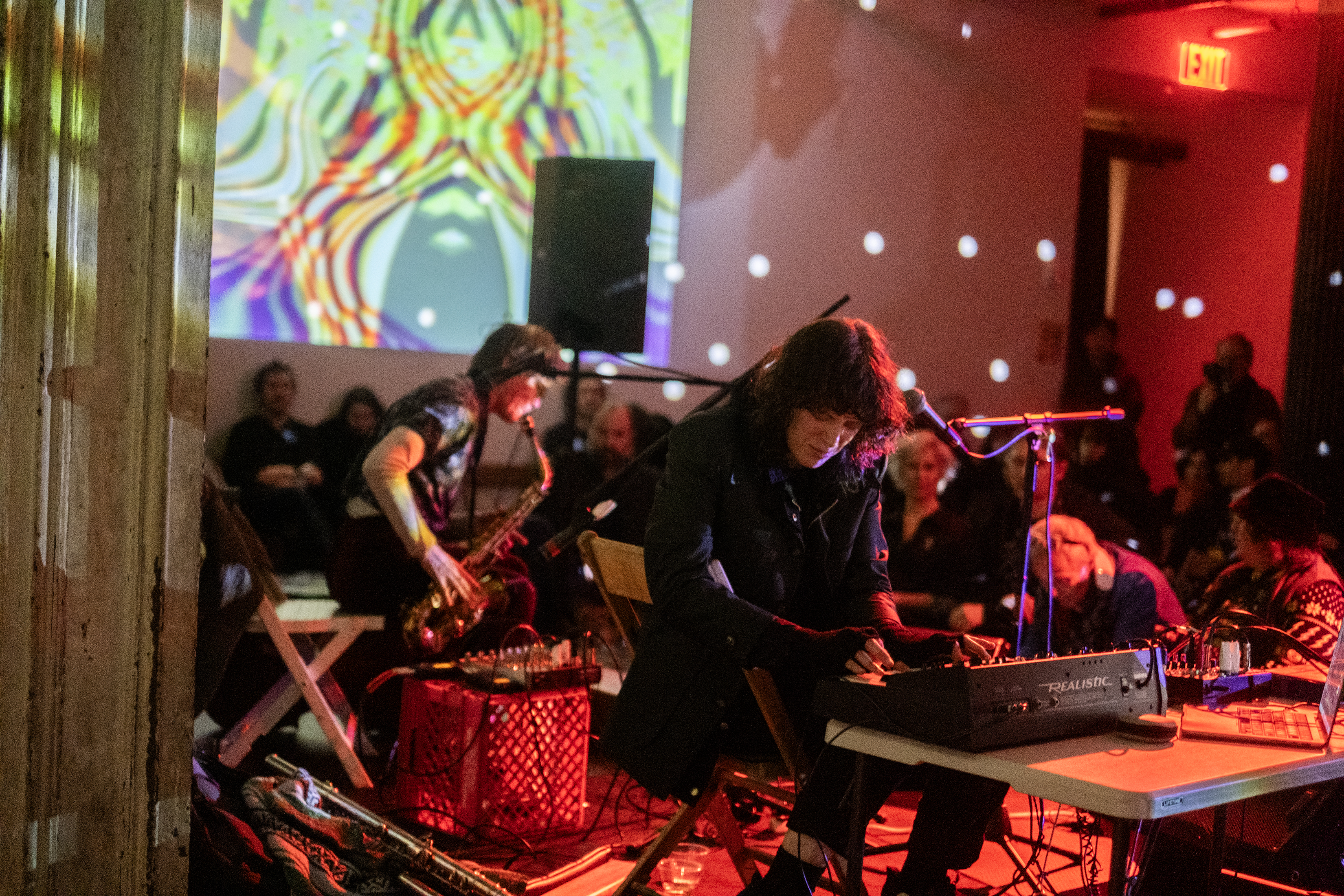 One performer operates a sound system, while the other plays the saxophone on stage in front of an audience. 