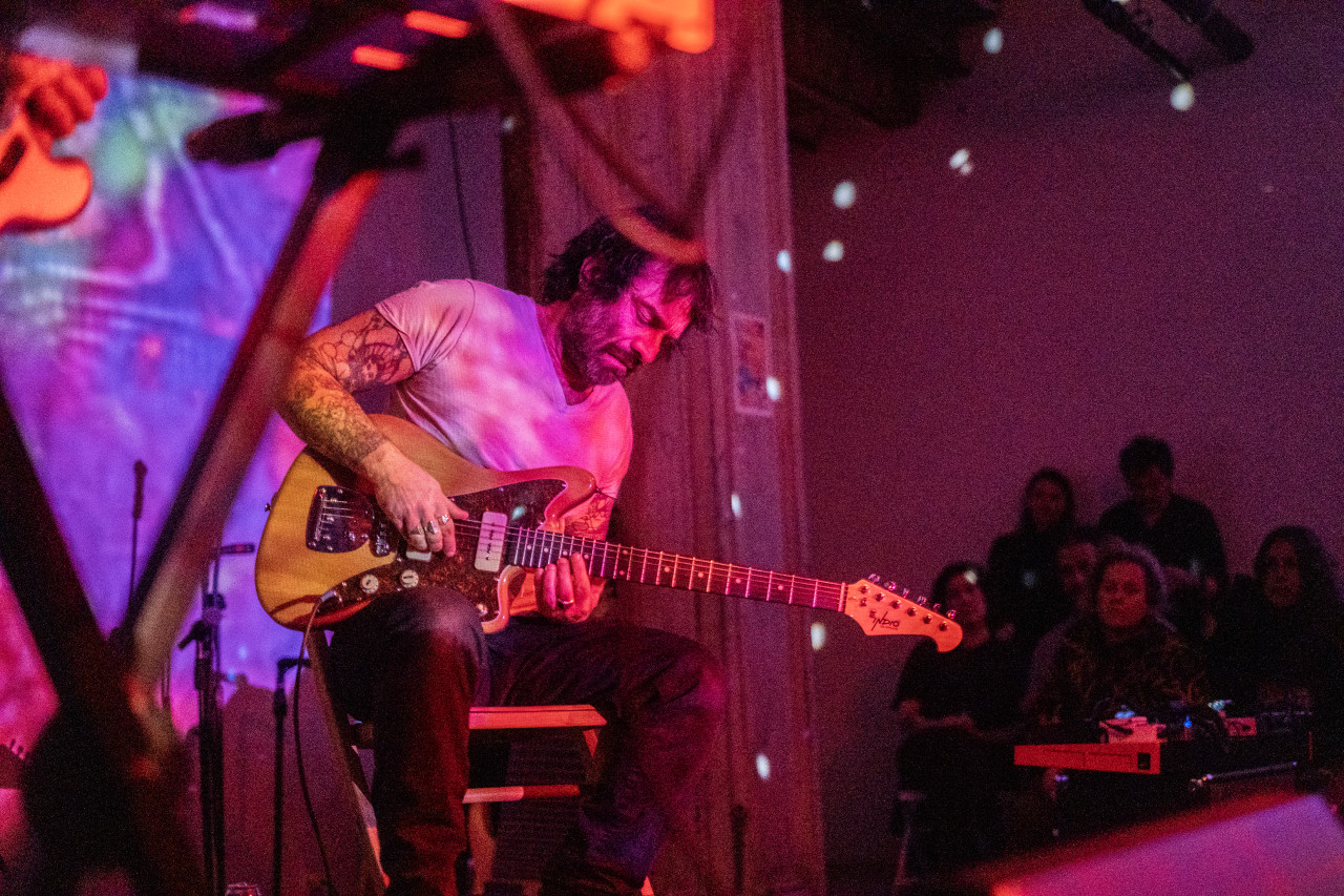 A seated performer plays the electric guitar on stage while bathed in pink light. Colorful patterns are projected onto the wall behind the stage.