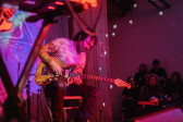 A seated performer plays the electric guitar on stage while bathed in pink light. Colorful patterns are projected onto the wall behind the stage. 