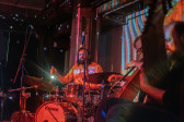 A performer plays the drums on stage, bathed in red light, while colorful geometric patterns are projected on the wall behind.