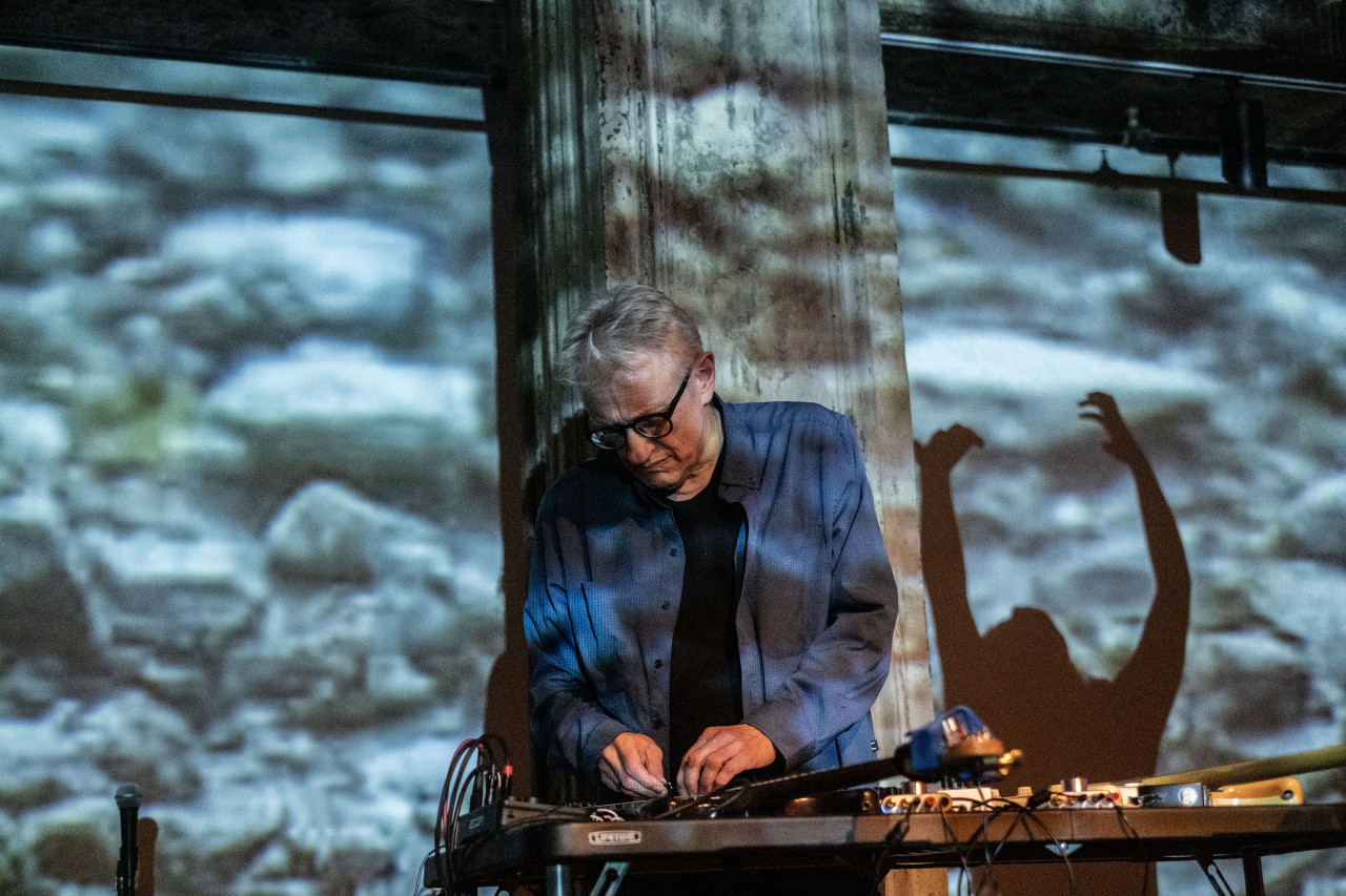 A performer operates a soundboard on stage. A shadow is cast on the wall behind, resembling another performer dancing with their hands thrown above their head.