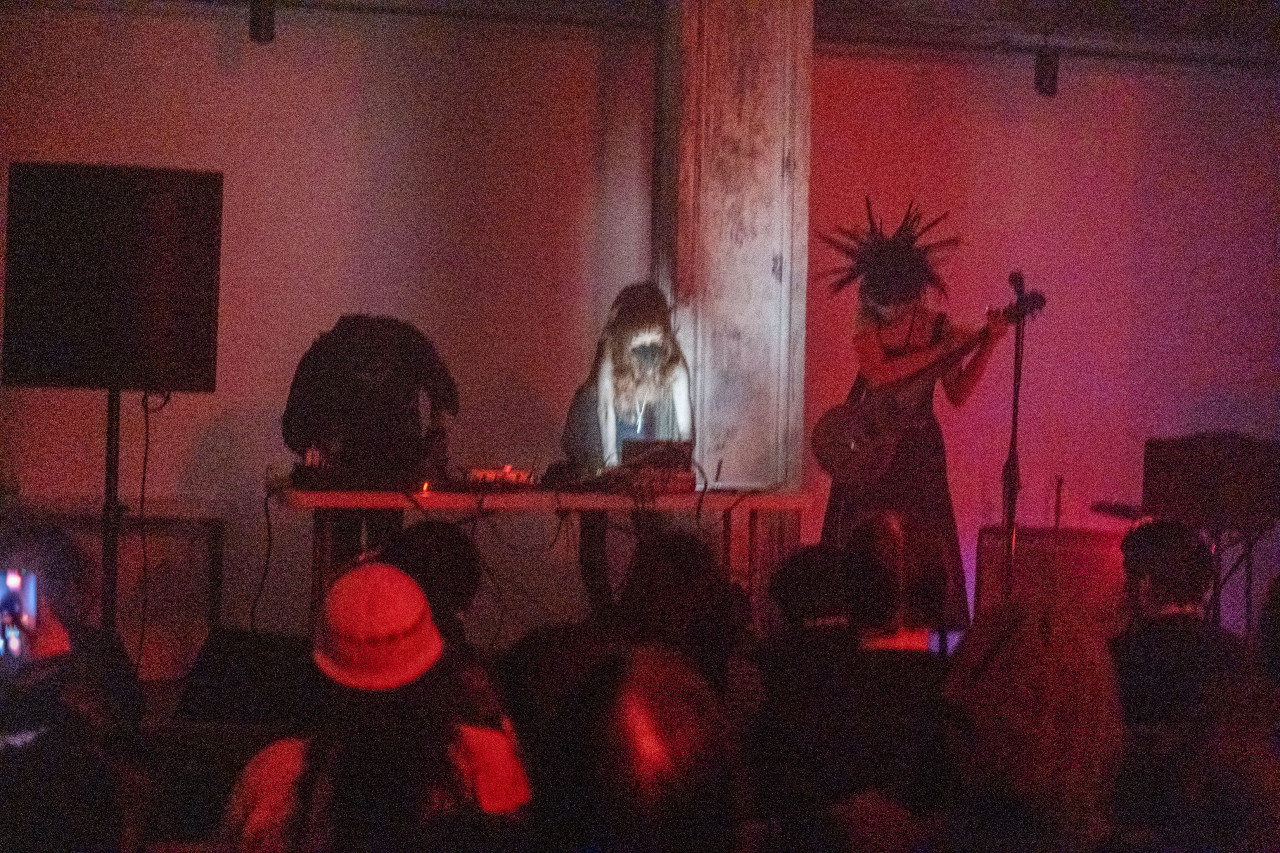 Three performers stand on stage in front of an audience.  Two performers operate at a table with musical equipment while the third plays an electric string instrument. The room is bathed in red light.