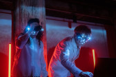 Two performers, wearing glasses adorned with bright, shining white lights, operate a soundboard. The stage is lit with two bright red tube lights. 