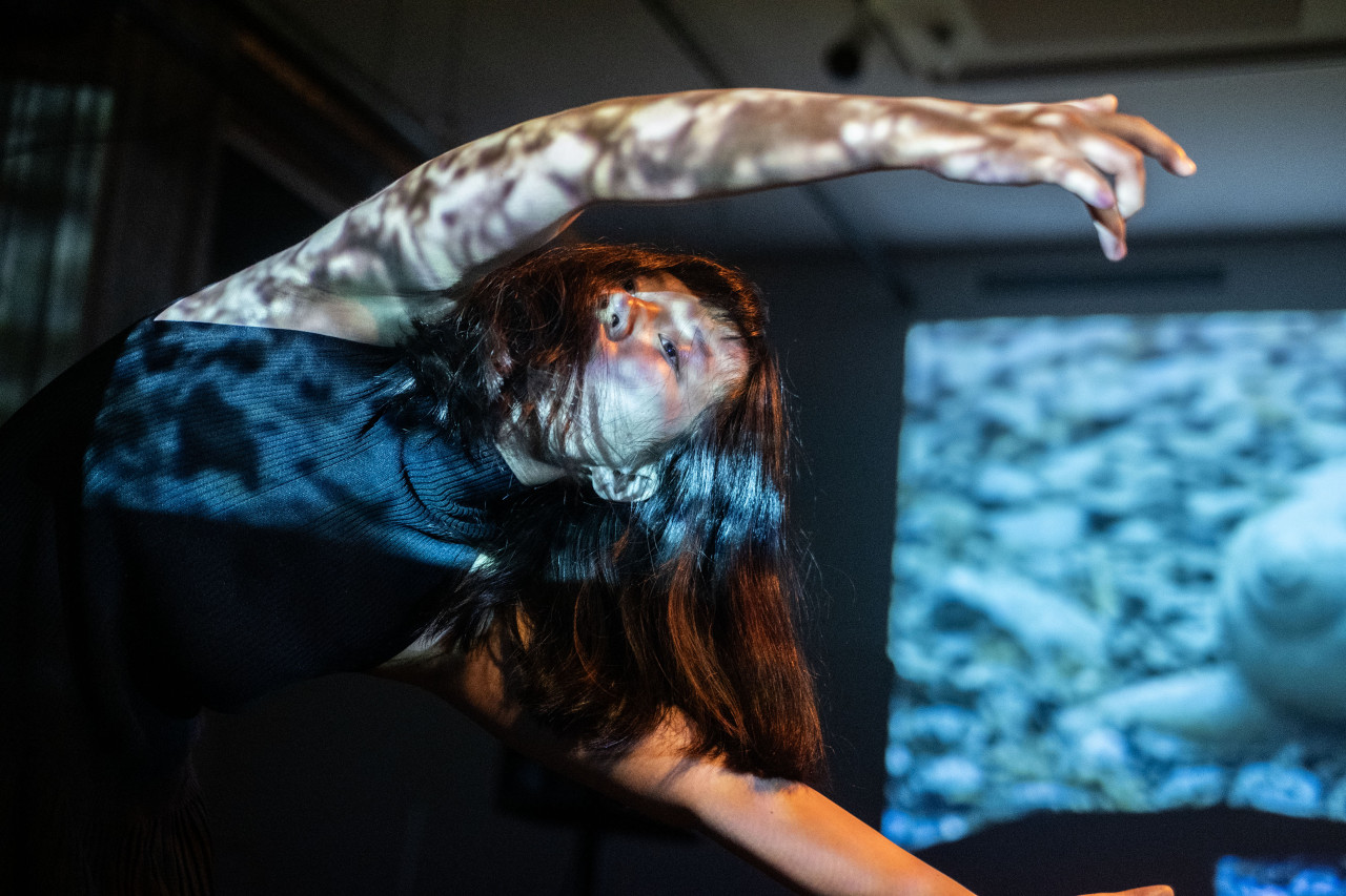 A performer caught in motion dances with their hands above their head, hanging at an angle. The performer is dappled in white lights.