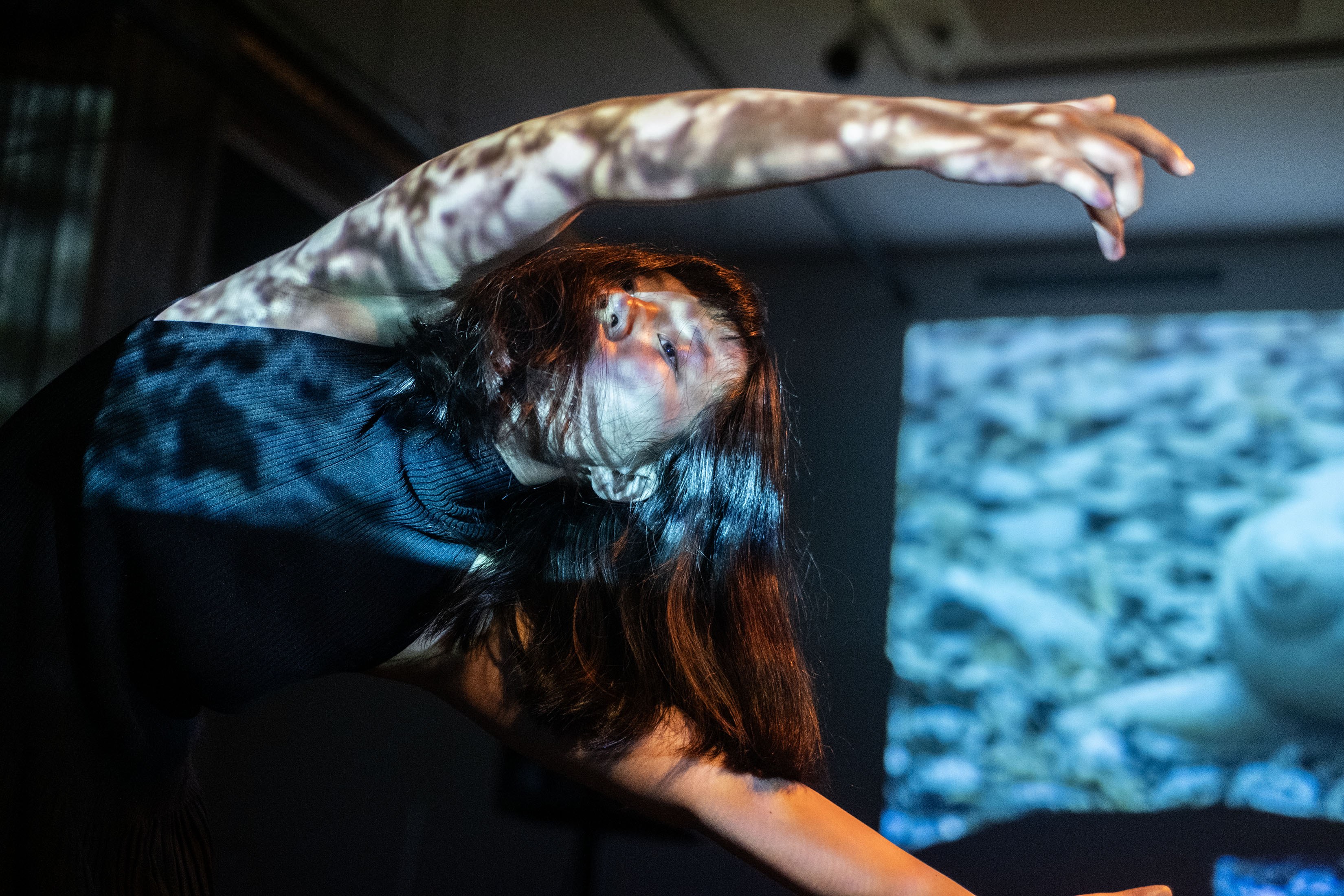 A performer caught in motion dances with their hands above their head, hanging at an angle. The performer is dappled in white lights. 