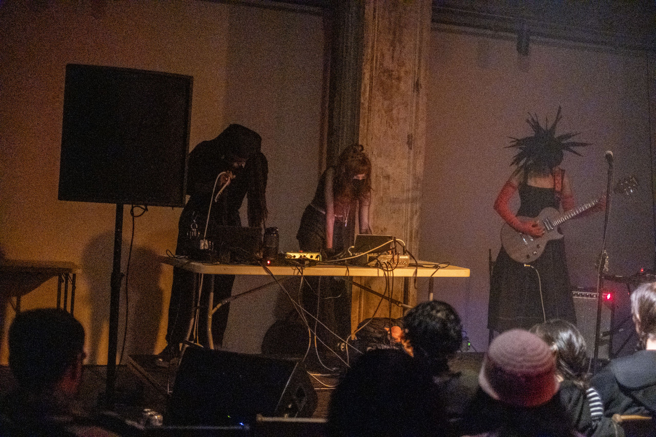 Three performers stand on stage in front of an audience.  Two performers operate at a table with musical equipment while the third plays an electric string instrument. The room is dimly lit.
