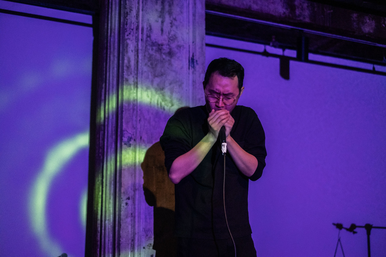 A performer sings into a microphone on stage. The performer and the wall behind the stage are bathed in purple light.