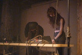 Two performers stand behind a table operating various musical equipment. 