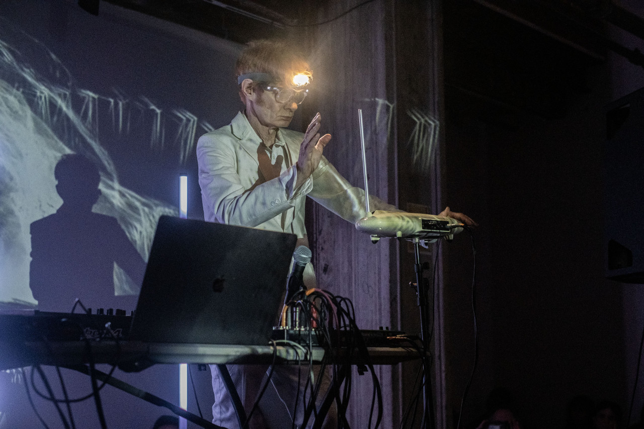 A performer, wearing a bright headlamp, plays a theremin on stage next to a table of musical equipment.